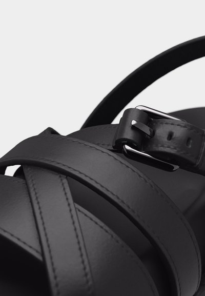 Κοντινό πλάνο μαύρων δερμάτινων λουριών σανδάλια με ασημί πόρπη σε ανοιχτό φόντο.