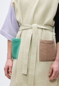 Μπέζ ρόμπα με υφή βάφλας και δεμένη ζώνη, με τσέπες σε τυρκουάζ και καφέ χρώμα, φορεμένη πάνω από πουκάμισο με μανίκια λεβάντας και μαύρου χρώματος.