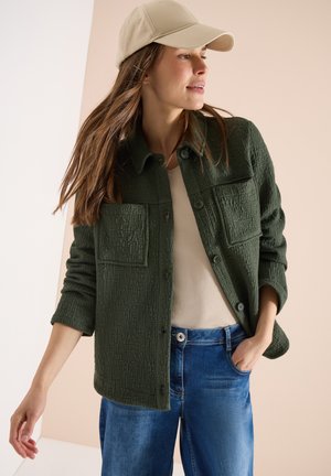 Kobieta w beżowej czapce, teksturowanej ciemnozielonej kurtce zapinanej na guziki, beżowym topie i niebieskich dżinsach, pozująca z jedną ręką w kieszeni i patrząca w prawo.