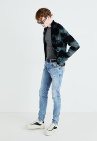 Cardigan noir à côtes avec motif tie-dye, chemise grise ajustée, jeans en denim léger bleu déchirés et baskets blanches avec accents noirs.