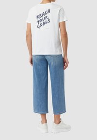 T-shirt en coton blanc avec le texte noir "ATTEINS TES OBJECTIFS" dans le dos. Associe à un jean large bleu clair et des baskets blanches.