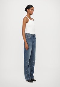 Lichtblauwe denim jeans met een losse pasvorm, voorzien van parelaccenten op de onderbenen en decoratieve patronen. Gecombineerd met een witte tanktop.