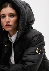 Černá péřová bunda s velkou kapucí, zdobená zlatým kováním a nášivkou s logem na rukávu. Hladký, lesklý povrch. Zapínání na zip vpředu.