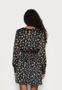 Kvinna med långt lockigt hår, iförd en svart blommig klänning med långa ärmar och en samlad midja, sedd bakifrån mot en enfärgad bakgrund.