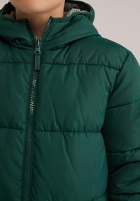 Zeleno podložena jakna z zadrgo, podloženo kapuco in subtilnimi šivi na prsih in ramenih. Gladek, vodoodporen material.