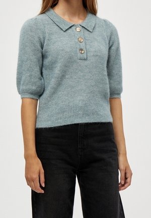 Woman wearing a light blue short-sleeve knit sweater with a collar and three buttons paired with black pants.