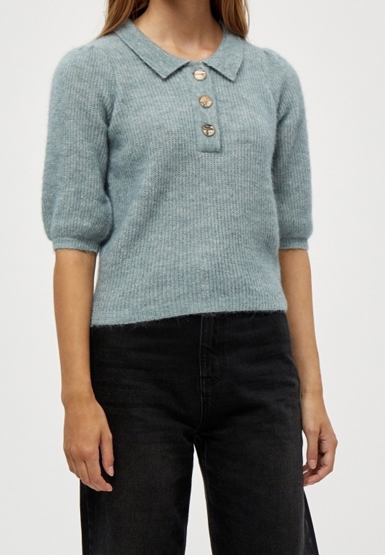 Femme portant un pull en maille bleu clair à manches courtes avec un col et trois boutons, associé à un pantalon noir.