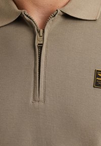 Beige polo-shirt met een gestructureerde brei, voorzien van een korte rits en een kraag. Inclusief een gouden rechthoekig logopatch op de voorkant.