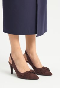 Brązowe zamszowe szpilki z odkrytą piętą, zakończone w szpic z kokardką z przodu, posiadające wąski obcas dla dodatkowej wysokości.