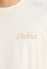Kreemivalge puuvillane T-särk, millel on ümar kaelus ja lühikesed varrukad, ning rinnal on peent beeži värvi teksti "Dickies".