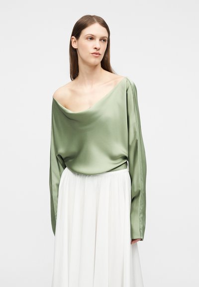 Νεαρή γυναίκα με μακριά καστανά μαλλιά, φορώντας μια άνετη πράσινη μπλούζα με ακάλυπτους ώμους και μια αέρινη λευκή φούστα, σε ουδέτερο φόντο.