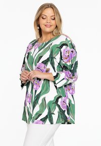Blumen-Tunika mit weißem Hintergrund, großen grünen Blättern und lila Tulpen. Aus weichem Stoff gefertigt, mit Dreiviertelärmeln und rundem Halsausschnitt.