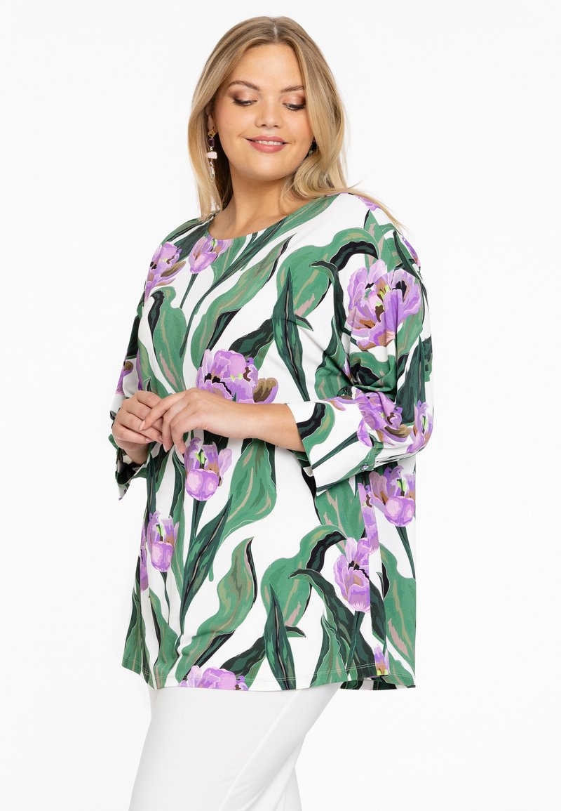 Blumen-Tunika mit weißem Hintergrund, großen grünen Blättern und lila Tulpen. Aus weichem Stoff gefertigt, mit Dreiviertelärmeln und rundem Halsausschnitt.