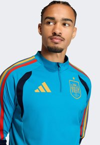 Vaaleansininen urheilupaita puolivetoketjulla, jossa laivastonsiniset yksityiskohdat ja puna-keltaiset raidat olkapäillä. Espanjan maajoukkueen logo näkyvissä.