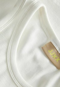 Bijela rebrasta tkanina meke teksture s blago zakrivljenim izrezom i oznakom koja prikazuje "JUXX" u žutoj boji na bež pozadini.