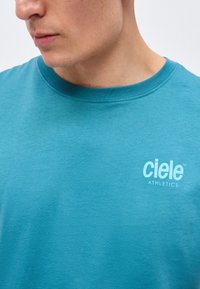 Detail osoby nosící modré tričko s kulatým výstřihem s logem "ciele ATHLETICS" vytištěným bíle na hrudi.