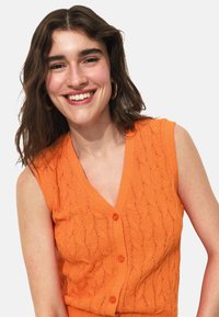 Gilet sans manches orange avec un col en V, motif en tricot torsadé texturé et cinq boutons sur le devant. Tissu en maille lisse.