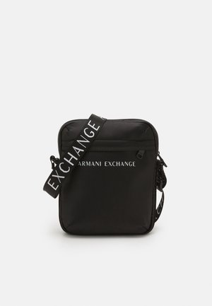 Kompaktowa czarna torebka na ramię z przednią kieszenią na suwak i regulowanym paskiem z napisem „EXCHANGE”, oznakowana logo "Armani Exchange" na biało.