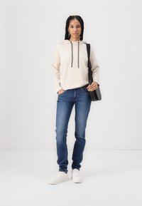 Sweatshirt bege com cordões, jeans azuis e ténis brancos. O modelo segura uma mala preta de ombro e está posicionado contra um fundo branco.