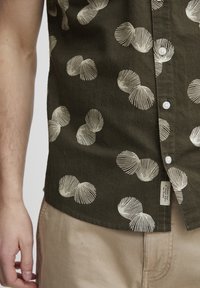 Camisa de manga corta color caqui con estampado de conchas blancas, cierre de botones en la parte delantera, cuello extendido y etiqueta en el dobladillo. Tela ligera con un acabado texturizado.