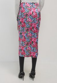 Femme portant une jupe crayon florale de longueur genou avec des fleurs roses, bleues et vertes, des collants noirs et des escarpins à bride arrière noirs, se tenant contre un mur blanc.