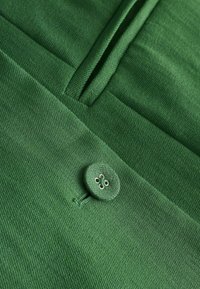 Gros plan d’un tissu vert avec un bouton recouvert du même tissu et un détail de poche fendue verticale sur un vêtement.
