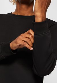 Felpa nera con colletto rotondo e polsini a costine, realizzata in un morbido tessuto textures. Persona che sistema la manica con una mano.