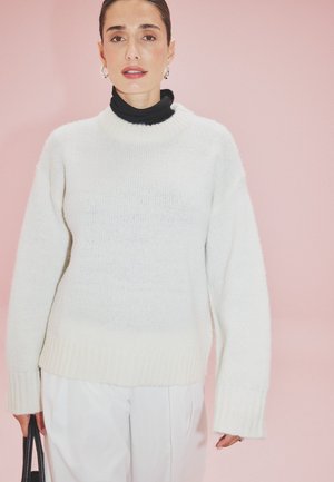 Maglione bianco a maglia con orlo a coste e vestibilità oversize, indossato sopra un dolcevita nero, abbinato a pantaloni bianchi. Sfondo rosa.