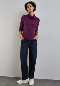 Maglione viola a collo alto con dettagli a bottone, abbinato a jeans a gamba larga blu scuro e sneakers bianche su una superficie testurizzata.