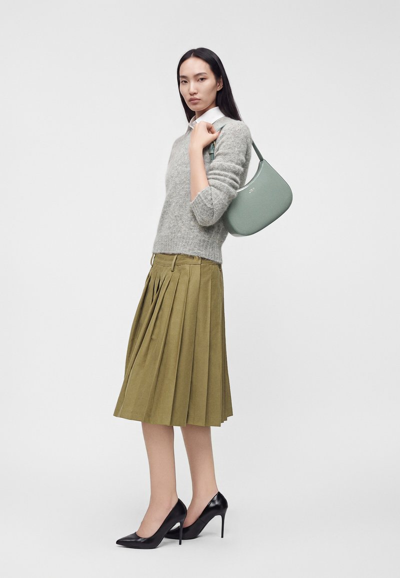 Femme portant un pull gris, une jupe plissée olive, des talons hauts noirs et portant un sac à épaule vert clair sur fond uni.