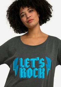 Šedé oversized tričko s modrým grafickým textem "LET'S ROCK" a akcenty blesků. Vyrobeno z měkké tkaniny, s širokým výstřihem.