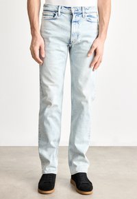 Svijetlo plave traperice opuštenog kroja, izblijedjeli izgled, pet džepova i suptilni detalji poput bora. Nošene s crnim antilop tenisicama.