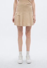 Beige geplooide mini-rok met een gladde textuur, een aangesloten tailleband en een rechte zoom. Gecombineerd met witte sneakers en geribbelde enkelsokken.