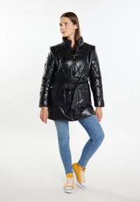 Cappotto piumino nero con vita cintata, zip frontale e due tasche anteriori. Abbinato a jeans azzurri e sneakers gialle. Materiale lucido.