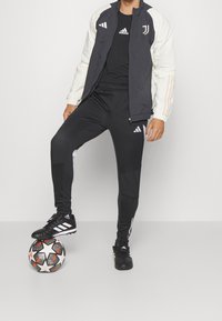 Schwarze und weiße Trainingsjacke mit Adidas-Logo, kombiniert mit schwarzen Hosen mit weißen Streifen und schwarzen Fußballschuhen auf einem Ball.