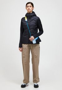 Black insulated jacket with a hood, chevron quilted pattern, paired with beige cargo pants and dark shoes, featuring a light blue accessory strap.