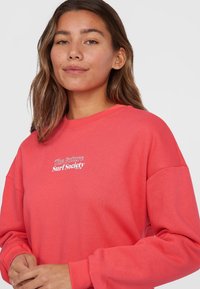 Korálová mikina s dlhými rukávmi, okrúhlym výstrihom a bielym nápisom "The Future Surf Society" na hrudi. Mäkko, zmes bavlny.