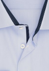 Lyseblå skjorte med nålestriber og krave med mørkeblå innerkant med hvide prikker, to hvide knapper synlige foran.