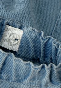 Svijetloplava traper tkanina s nabranim elastičnim pojasom i prozirnim gumbom pričvršćenim za bijelu tekstilnu oznaku unutar odjeće.