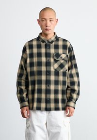Camisa de franela a cuadros en negro y beige. Presenta un cierre de botones en el frente, cuello puntiagudo y un bolsillo en el pecho. La tela parece texturizada y cálida.
