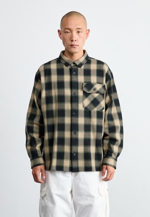 Camisa de franela a cuadros en negro y beige. Presenta un cierre de botones en el frente, cuello puntiagudo y un bolsillo en el pecho. La tela parece texturizada y cálida.