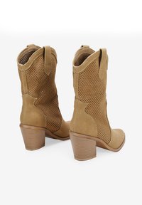 Botas tipo botín en ante color marrón con diseño perforado, puntera redondeada y tacón apilado. Con tiradores a cada lado para una fácil puesta.