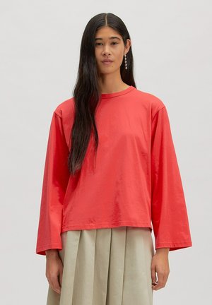 Vrouw met lang donker haar die een losse rode top met lange mouwen en een beige geplooide rok draagt, staand tegen een effen lichte achtergrond.