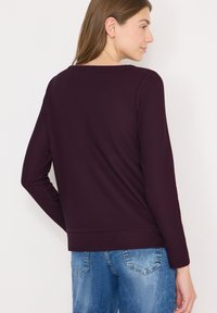 Viininpunainen pitkähihoinen toppi jossa on teksturoitu ribbineuloskuvio, pyöreä kaula-aukko ja vartalonmyötäinen vyötärönauha, yhdistetty sinisiin farkkuihin.