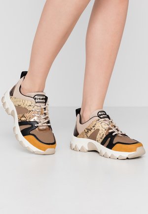 Jambes portant des baskets beiges, marron et noires avec des semelles blanches dentelées et le logo "BUGATTI" sur la languette, sur un sol et un arrière-plan gris clair.