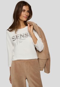 Junge Frau trägt ein weißes Langarmshirt mit dem Schriftzug "SENSE", beige Cordhosen und hält eine passende beige Jacke über der Schulter.