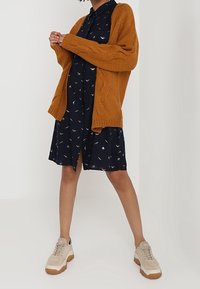 Cardigan lavorato a maglia color ruggine sopra un vestito blu navy con piccoli stampati di uccellini, abbinato a sneakers beige con suola spessa.