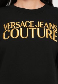 Čierna mikina s veľkým zlatým vyšívaním "VERSACE JEANS COUTURE" cez hruď, s okrúhlym výstrihom a rebrovanou textúrou.