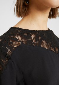 Kvinna som bär en svart topp med blommig spetsdetalj på axlarna och övre bröstet, med kort brunt hår och mörka, runda örhängen.