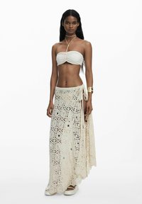 Krémová maxi sukňa z háčkovanej čipky s precízne zdobenými šnúrkami a bočným viazaním. Spárovaná s jednoduchým bandeau topom a bielymi platformami.
